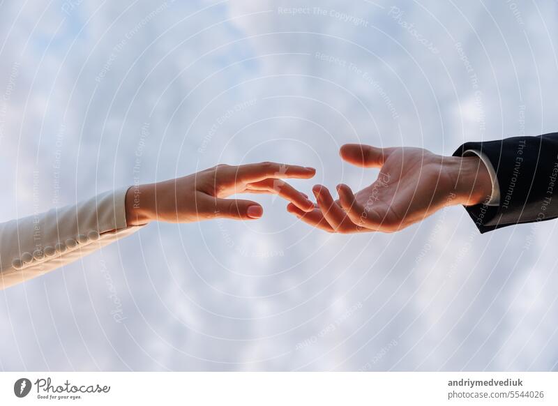 Hands of bride in white dress and groom in suit reaching each other, touching fingers on blue sky background. Helping hands for save and support people concept. Wedding day. Valentine day.