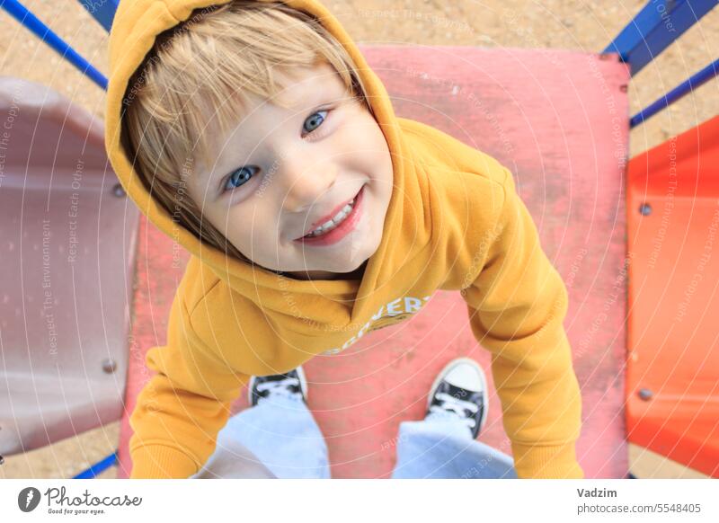 A cheerful child in a yellow sweater plays with his mother on the playground, holds her legs and looks up at her. Family people children Caucasian walk