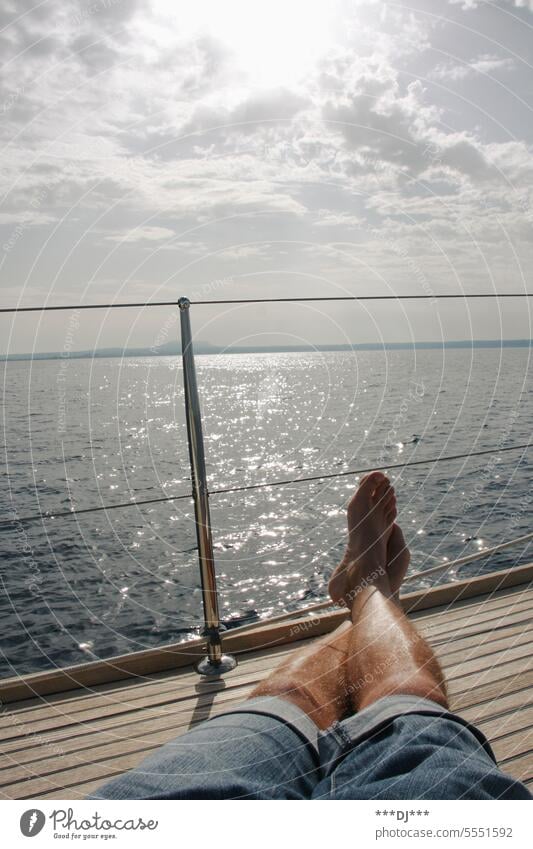 Man with short jeans trousers sitting or lying on a boat deck with outstretched, crossed legs - direction of view out to the wide sea. Legs feet Human being