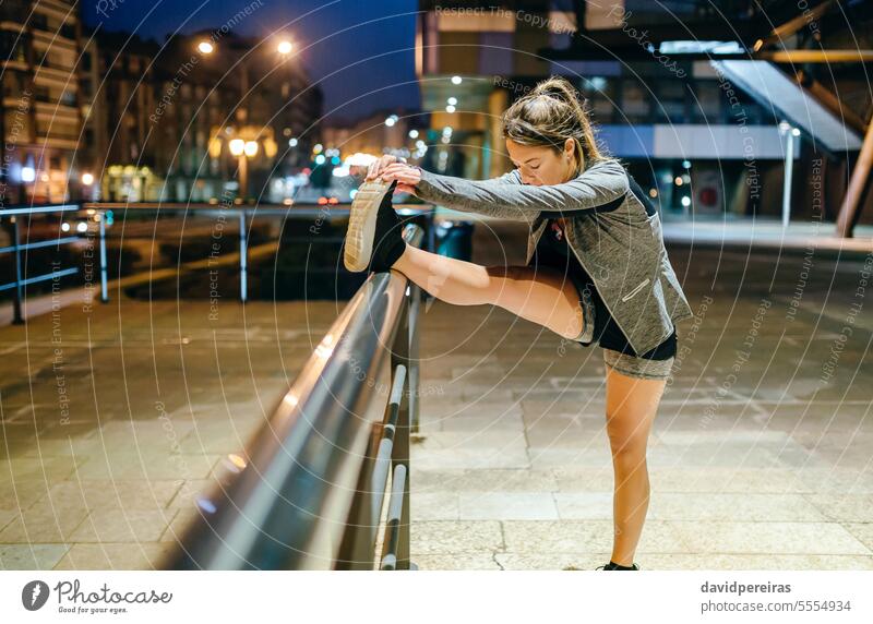 Woman runner stretching her leg before training at night on town female woman warming up exercise evening banister railing blonde young ponytail outside urban