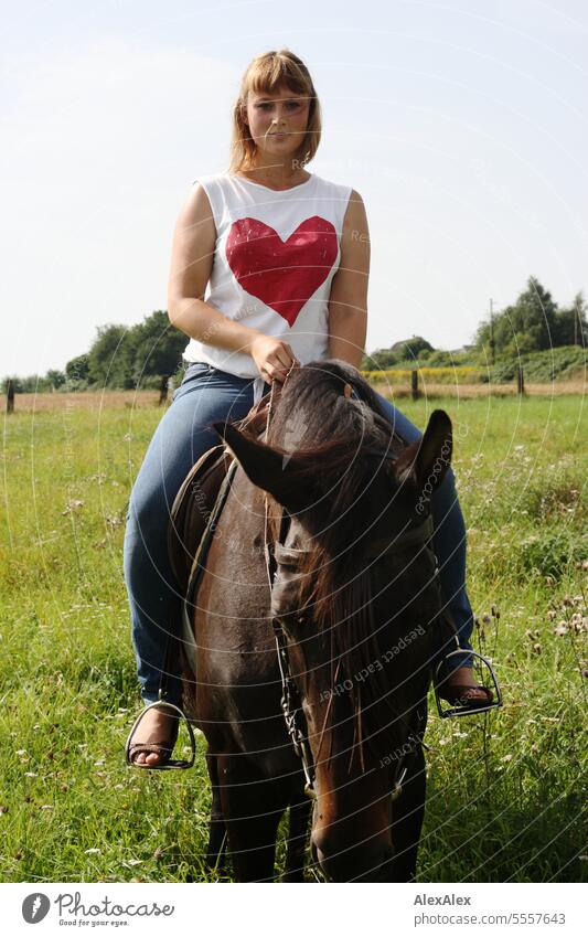 Portrait of young woman with big heart on t-shirt riding brown horse in green pasture Woman Young woman Dress Garden Slim Athletic pretty attractive lovely