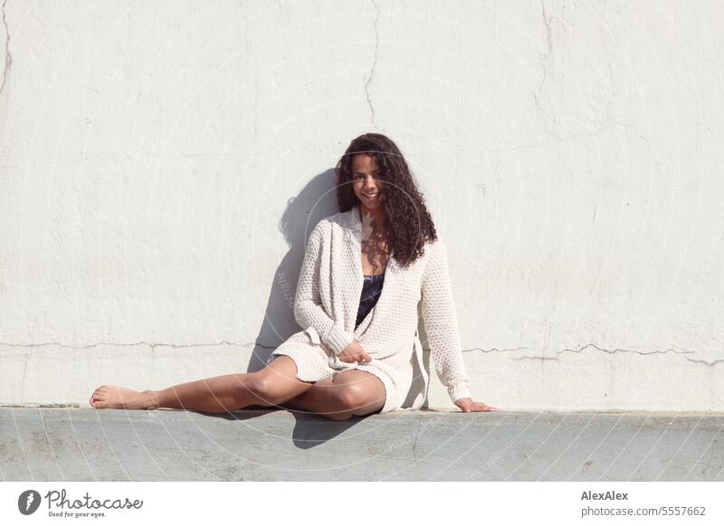 A young beautiful woman sits on a threshold of a concrete wall and smiles Woman Young woman Long-haired Curly pretty Athletic Slim Legs Barefoot attractive