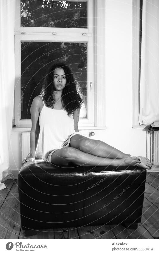 A young beautiful woman sits on a square sofa block in a room and looks into the camera - black and white shot Woman Young woman Long-haired Curly pretty