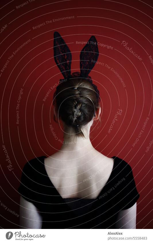 Portrait of young woman from behind in front of red wall wearing black lace bunny ears and black top - dark sexy easter image Woman Young woman Back portrait