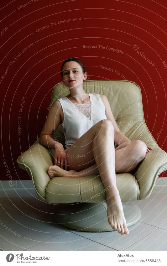 Young, slim, athletic woman sits in a beige leather armchair in front of a red wall and poses for the camera Woman Young woman Slim Athletic pretty fit Legs