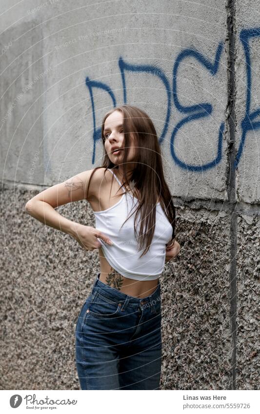 A stunning brunette woman, dressed casually in blue jeans, exudes a sense of wild freedom as she strikes a natural pose beside a suburban mural and gray walls. An inked beauty, immortalized in a captivating portrait.