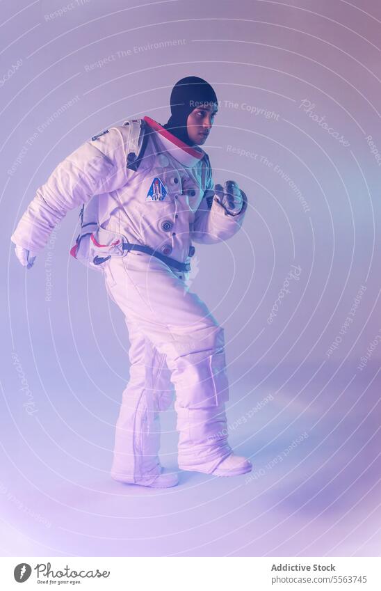 Male ethnic astronaut in spacesuit and backpack with dark hood marching against purple background cosmonaut explore cosmos astronomy mission gadget planet man