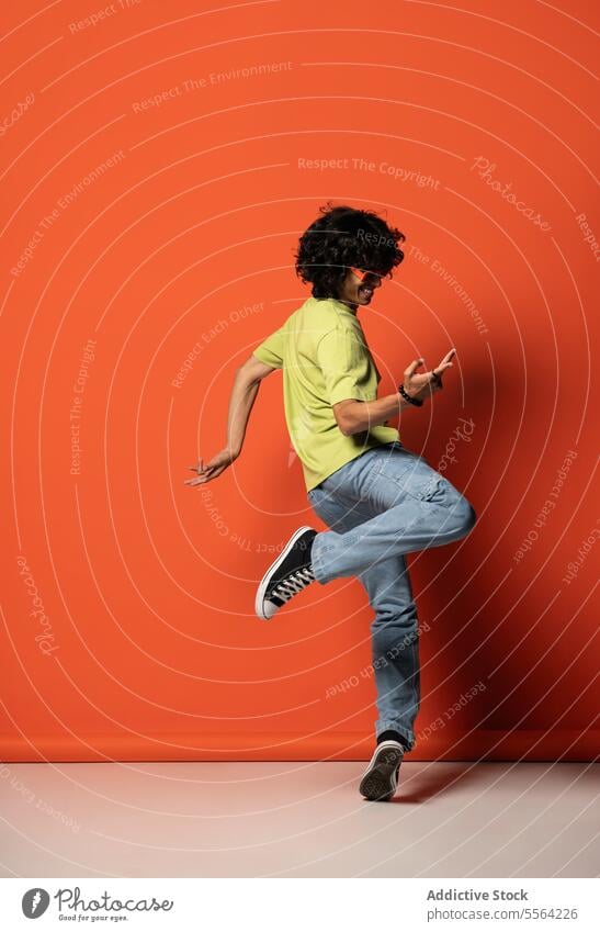 Young man in dark hair dancing near red wall dance leg raised dancer shadow perform move flexible light energy male casual sneakers arm raised bead bracelet