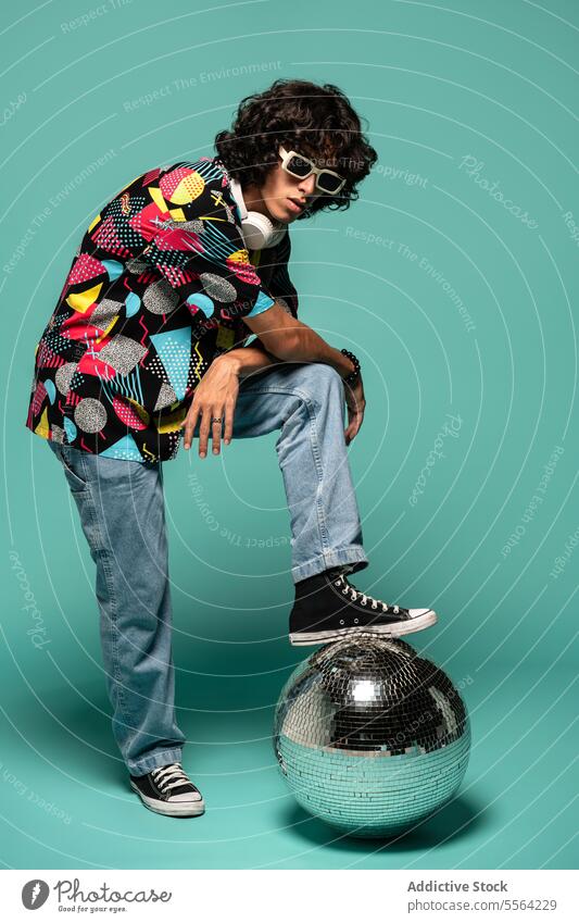 Stylish ethnic young man in colorful shirt standing with leg on disco ball near turquoise backdrop sunglasses lean on knee funky fashion leg raised male