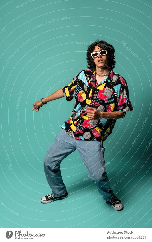 Stylish young ethnic man dancing with moving legs and arms near turquoise wall funky dancer perform excited move energy sunglasses male sit latin american