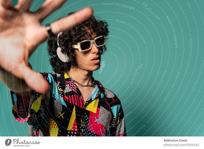 Trendy ethnic young man with headphones and sunglasses near turquoise backdrop show colorful gesture shirt funky fashion listen male stand latin american