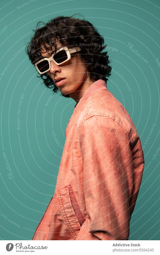 Confident young ethnic man in sunglasses pink jacket standing near turquoise backdrop funky dancer fashion curly hair cool individuality male latin american