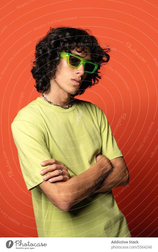 Confident young ethnic man in sunglasses standing near turquoise backdrop funky dancer fashion curly hair cool individuality male latin american arms crossed