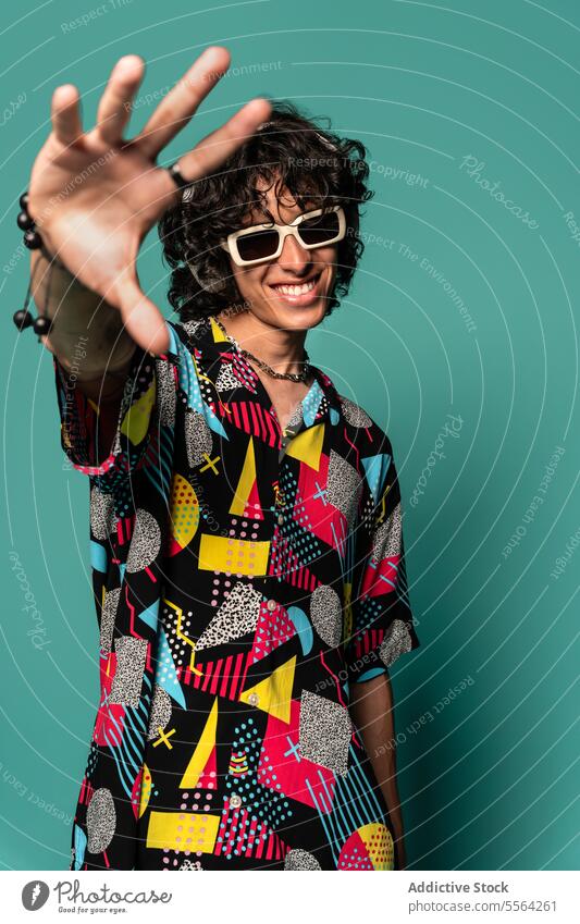 Trendy ethnic young man with headphones and sunglasses near turquoise backdrop show colorful smile gesture shirt funky fashion listen male latin american