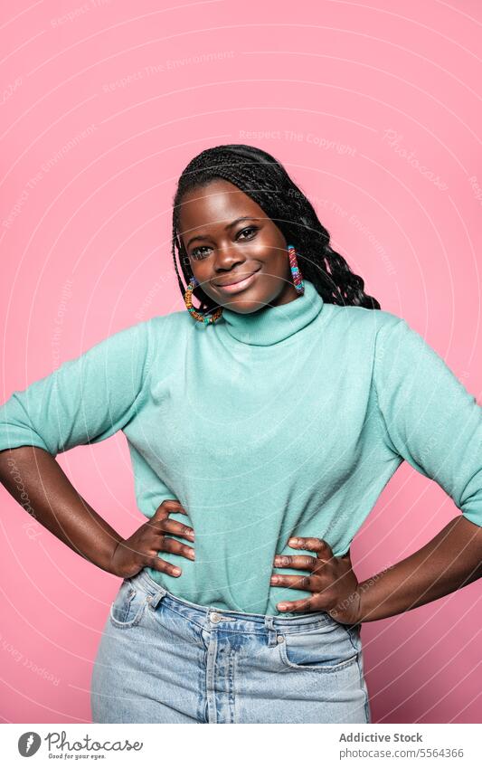 Smiling African woman with hands on hips smiling african braided hair colorful earrings turquoise turtleneck pink background happiness pose confidence beauty