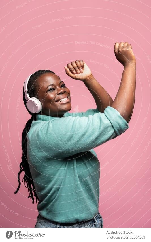 Joyful african woman with headphones pink background joy music listen raise hand smile youth emotion happiness celebrate dance technology audio melody rhythm