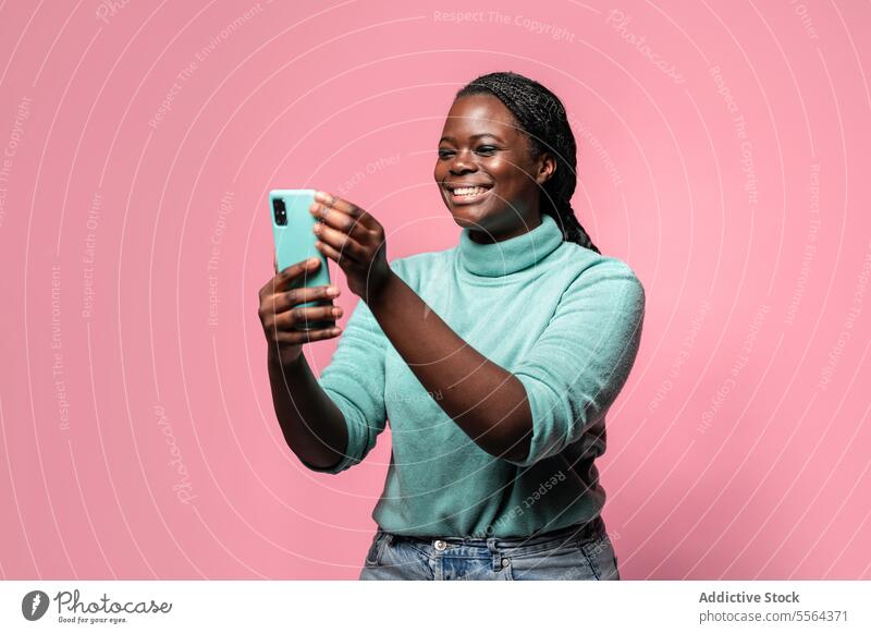 Cheerful african woman using phone against pink background smartphone turquoise turtleneck teal capturing moment backdrop joyful smile technology mobile photo