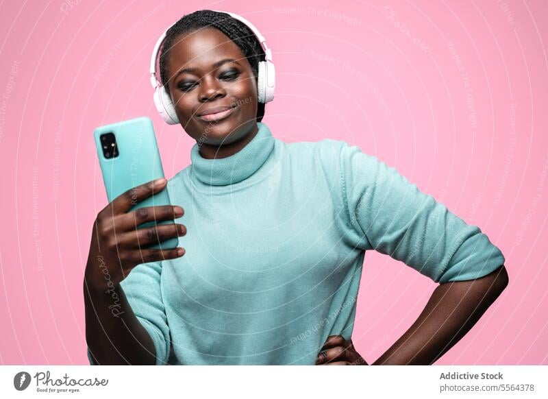 African woman taking selfie with smartphone african turquoise sweater mint green pink headphones backdrop pose capture photo mobile device fashion technology
