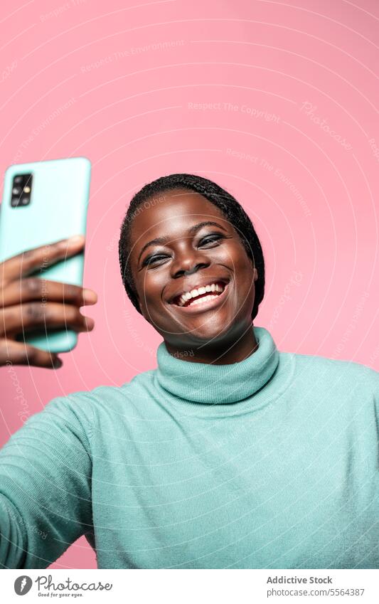 Cheerful african woman taking a selfie against pink background close-up face smile smartphone teal emotion joy eyes makeup lashes teeth laugh portrait photo