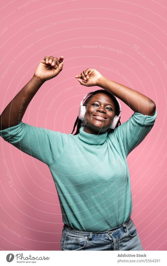 Joyful African woman with headphones african pink background joy arms raised dancing music happiness green top entertainment celebration audio mood lively