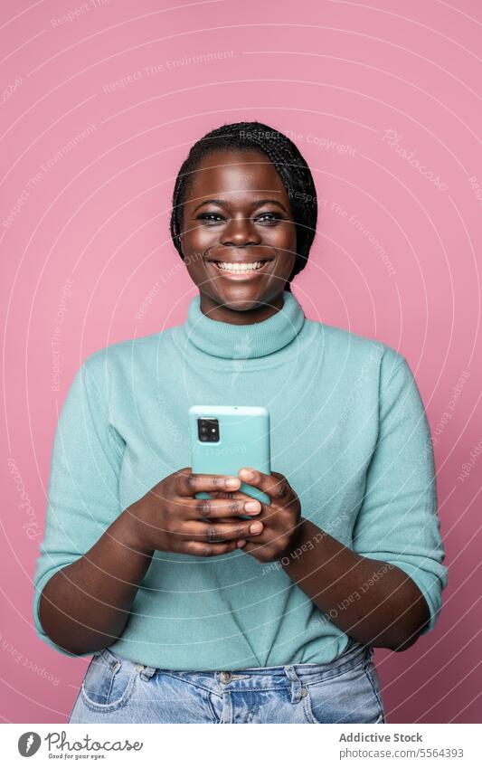 African joyful woman with smartphone and blue sweater against a pink background cheerful turquoise turtleneck light blue smile broad young teeth braids fashion