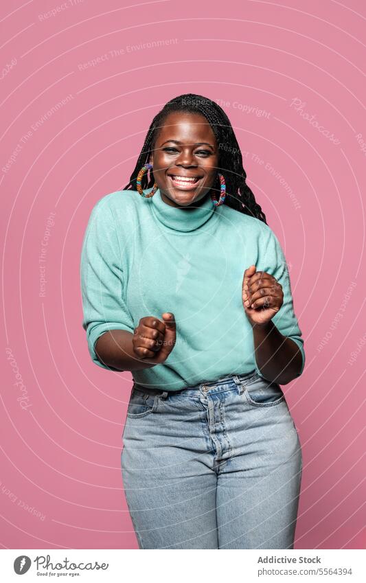Joyful African woman in turquoise sweater african dance jeans movement pink background smile joy excitement fist clench denim fashion pose portrait earring