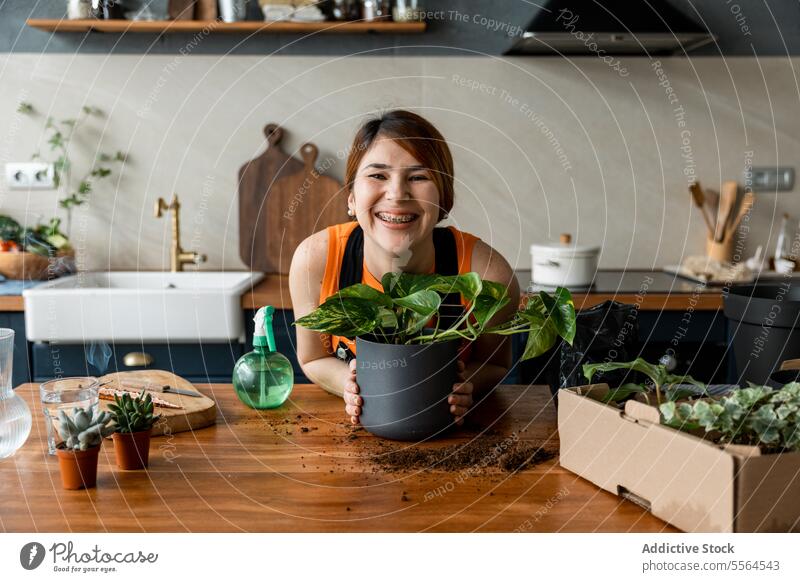 Cheerful young woman holding pot with green plant during home gardening gardener apron surface box smile at home female stand lean on wooden happy work hobby
