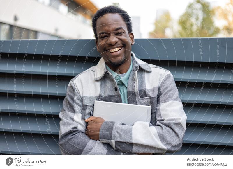 Cheerful young black man standing with laptop in park in daytime smile portrait happy cheerful building positive city arms crossed summer male african american