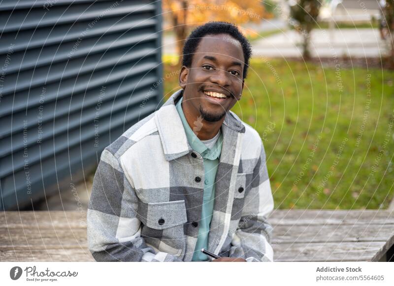 Cheerful young black man sitting on bench with pen and pen in daytime smile portrait happy positive building sidewalk male ethnic african american city cheerful