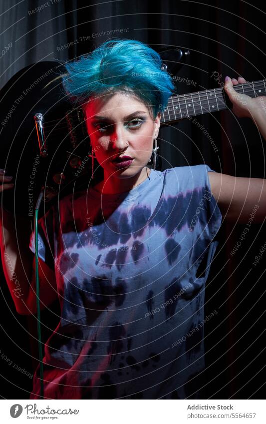 Young stylish woman carrying electric guitar behind head in dark room musician serious guitarist instrument perform stage portrait dyed hair young female makeup