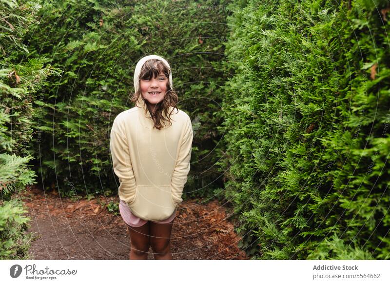 Smiling preteen girl in hoodie standing near green trees kid coniferous park road happy smile countryside child hand in pocket positive nature maze content
