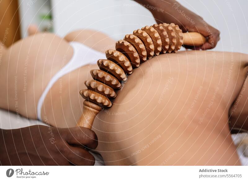 Crop masseur massaging back of unrecognizable female customer with wooden instrument in salon massage shoulder roller procedure treat therapy wellness therapist