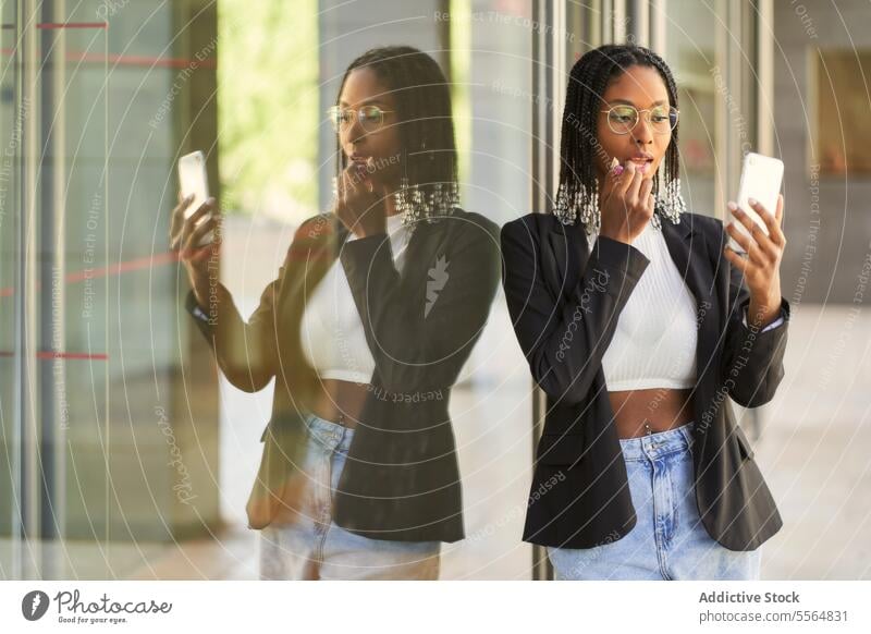 Black businesswoman using smartphone as mirror doing makeup lipstick entrepreneur reflection braids hairstyle window confident glass focus black female modern