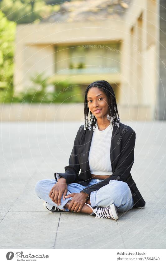Smiling African American female sitting on floor in yard of building woman smile appearance confident casual portrait happy style afro positive braid pleasant