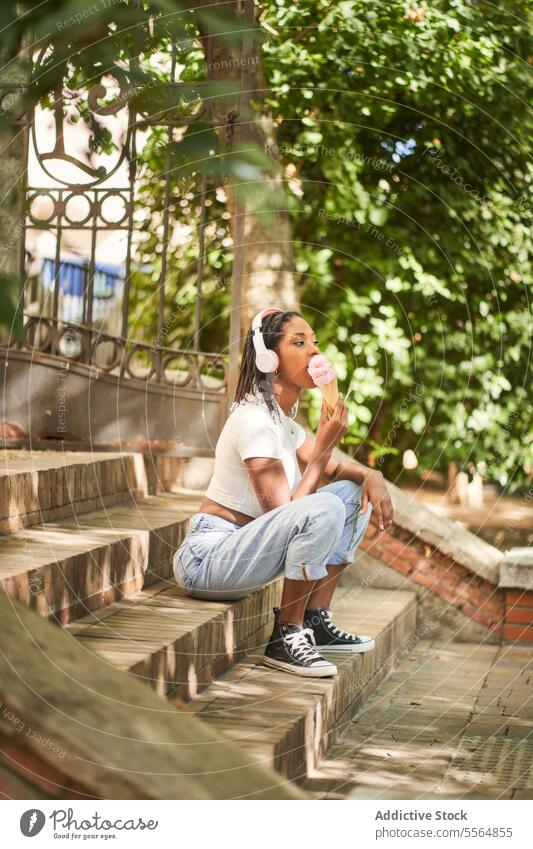Black woman student in headphones and with ice cream cone sitting on stair spoon campus stairway summer female young ethnic black african american gadget device