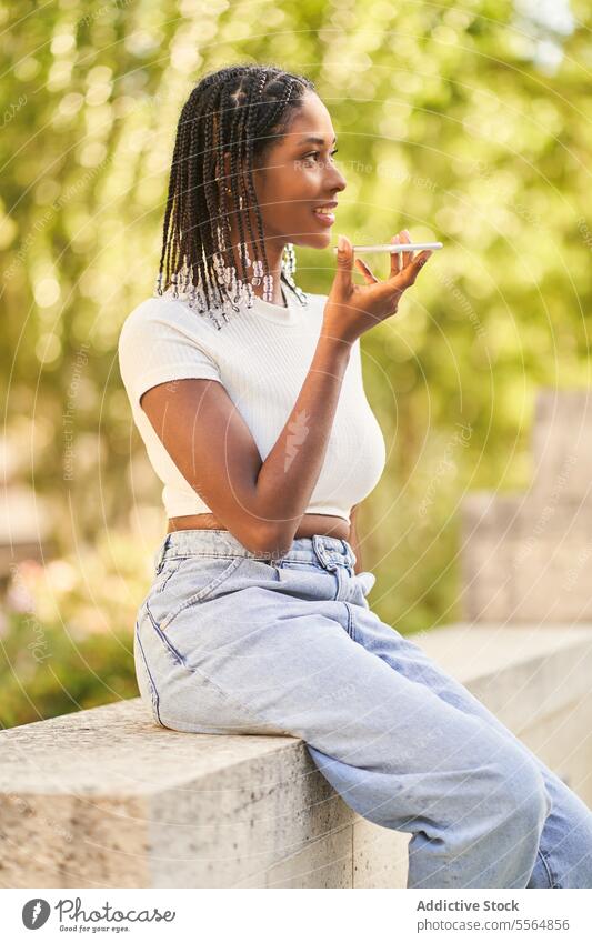 African American woman sitting on stone bench and recording voice message using smartphone audio border park cellphone connection afro hairstyle braid app