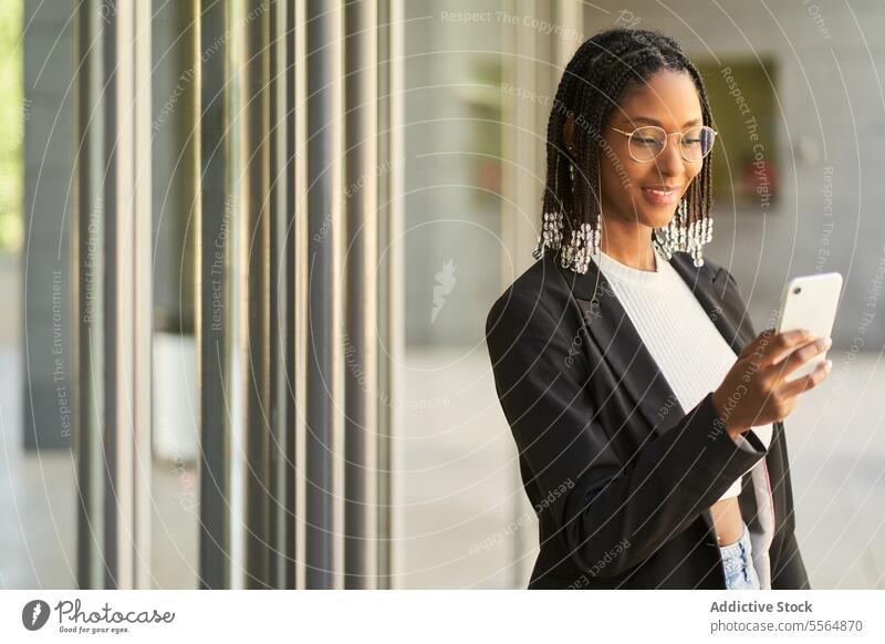 Smiling businesswoman standing and using mobile phone while holding laptop smartphone browsing entrance smile entrepreneur modern office doorway gadget