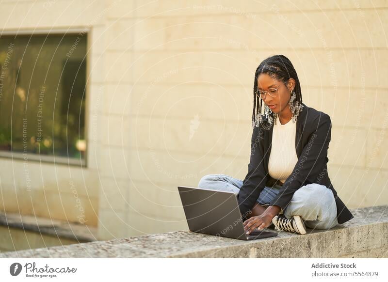 Focused ethnic businesswoman working on laptop while sitting at stone border near plants freelance using remote pc browsing project internet netbook online