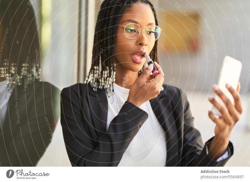 Black businesswoman using smartphone as mirror doing makeup lipstick entrepreneur reflection braids hairstyle window confident glass focus black female modern