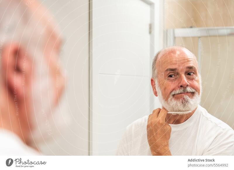 Senior man shaving beard with blade shave mirror at home morning routine bathroom reflection positive smile male senior aged elderly gray hair care hygiene