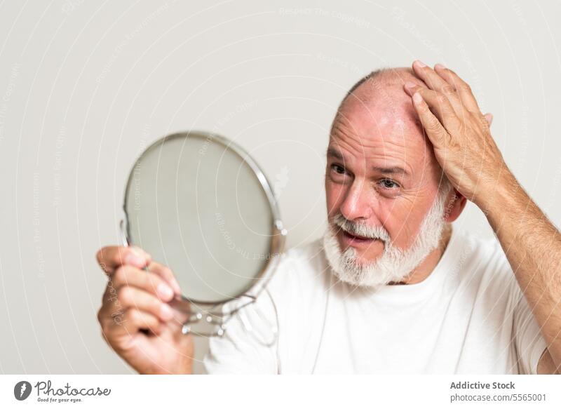 Senior bald male model looking at mirror in studio man wrinkle hairless appearance complexion grooming self care routine daily beard senior gray hair procedure