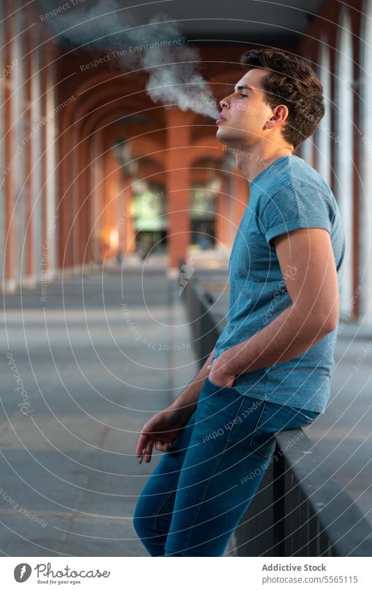 Young man exhaling smoke near arches in urban place. caucasian Gen-Z railing young leaning jeans shirt outdoor building evening light profile casual city