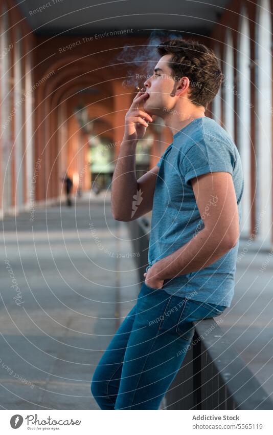 Young man smoking in city sitting in urban railing. young exhale smoke Gen-Z standing blue t-shirt jeans caucasian sidewalk brunette hair cigarette male