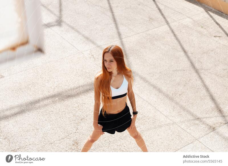 Young redhead woman stretching on sunlit street outdoors sunny day sports bra black shorts pavement fitness exercise active health sunlight long hair focus