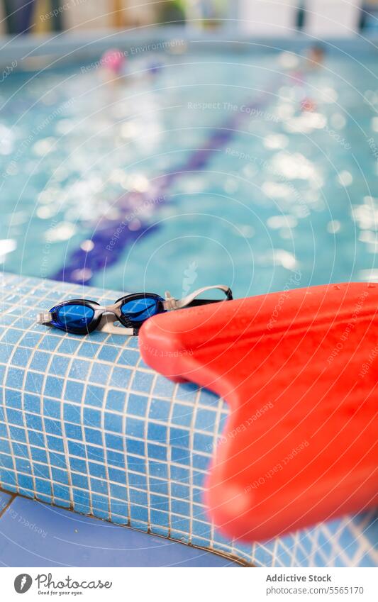 Swim goggles and kickboard at pool edge with blurred child indoor blue tile swim water equipment sport training recreation facility leisure swimmer rest