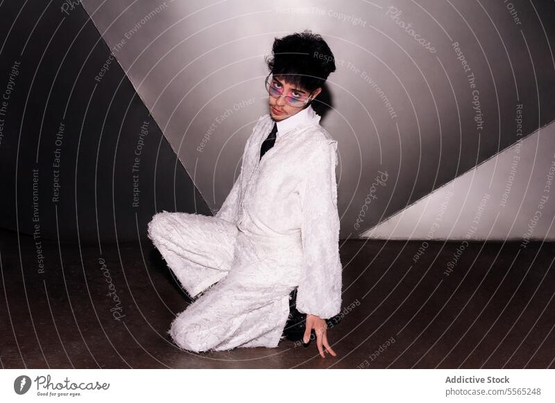 Man Daring gaze in a radiant suit. individual makeup sitting concrete floor white camera dual-tone wall fashion-forward strong trend edgy style glasses tie lace