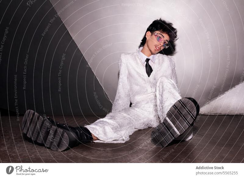 man Youthful expression with edgy style. makeup individual reclining concrete floor white suit lustrous chunky boots two-toned backdrop youthful fashion pose