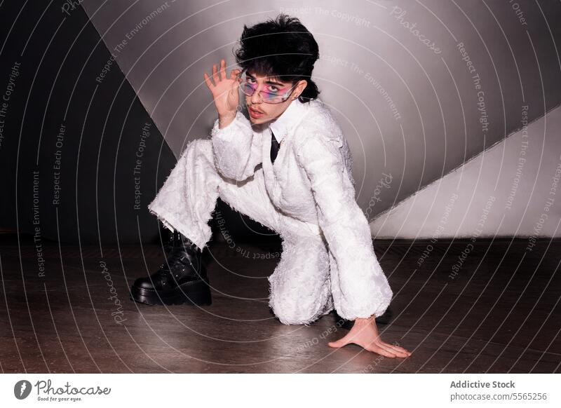 Man Daring gaze in a radiant suit. individual makeup sitting concrete floor white camera dual-tone wall fashion-forward strong trend edgy style glasses tie lace