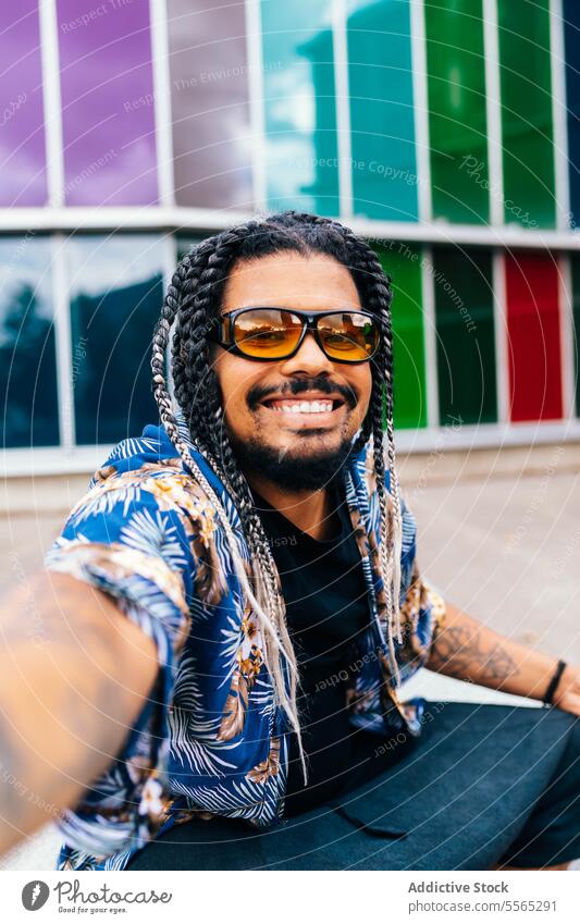 Smiling latin man taking a selfie in front of multicolored glass panels. braids sunglasses smiling close-up vertical colorful background eyes joy happiness