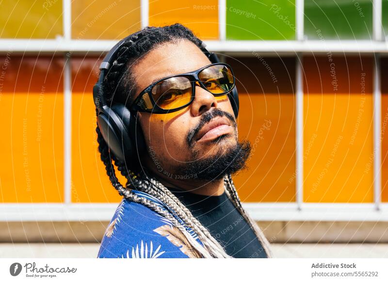 Portrait of a latin man with headphones and sunglasses in front of building colorful panels. braid aesthetic beard close-up portrait outdoor profile face